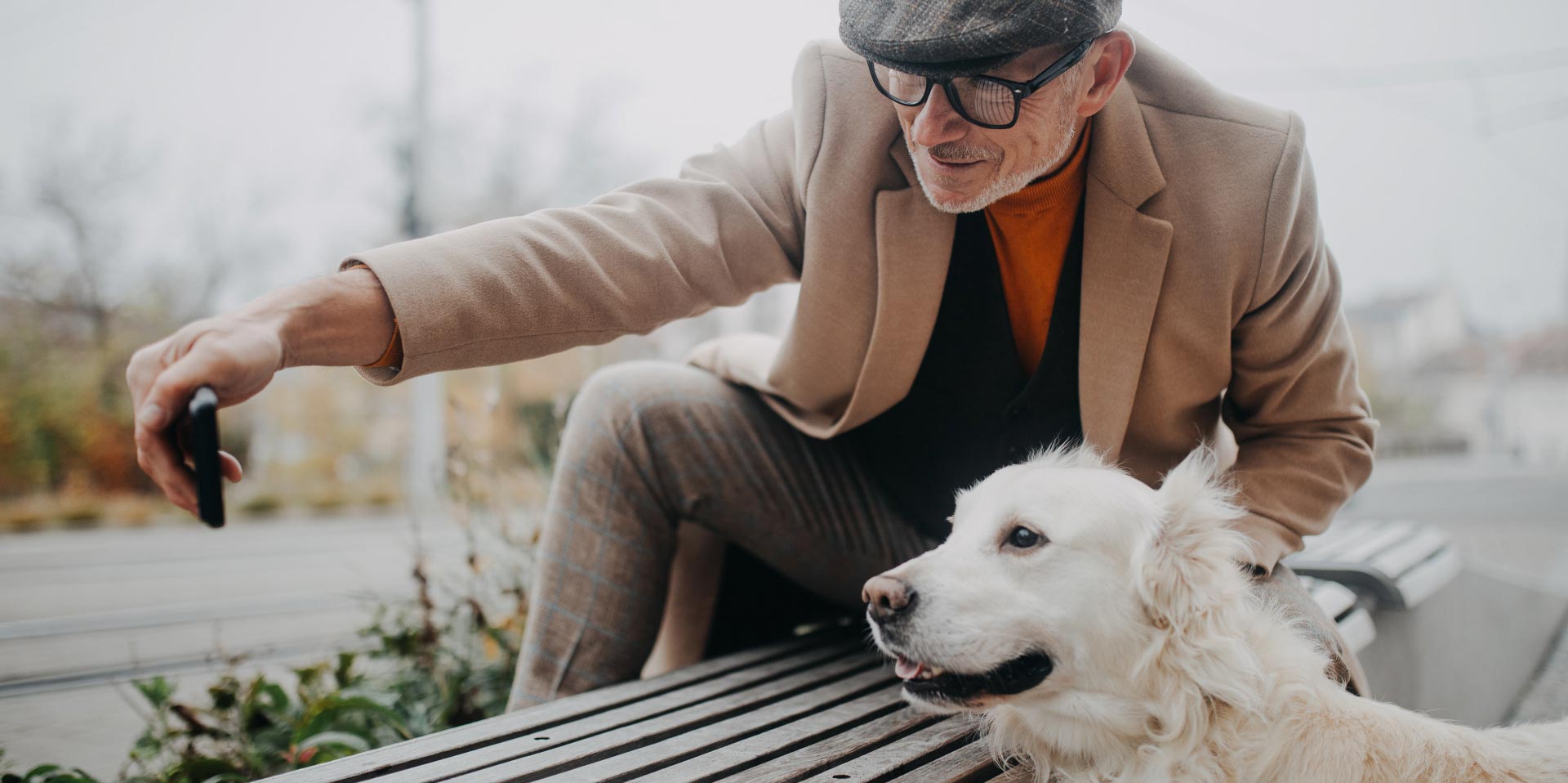 Velkledd mann som tar bilde av seg selv og sin hund på en benk.