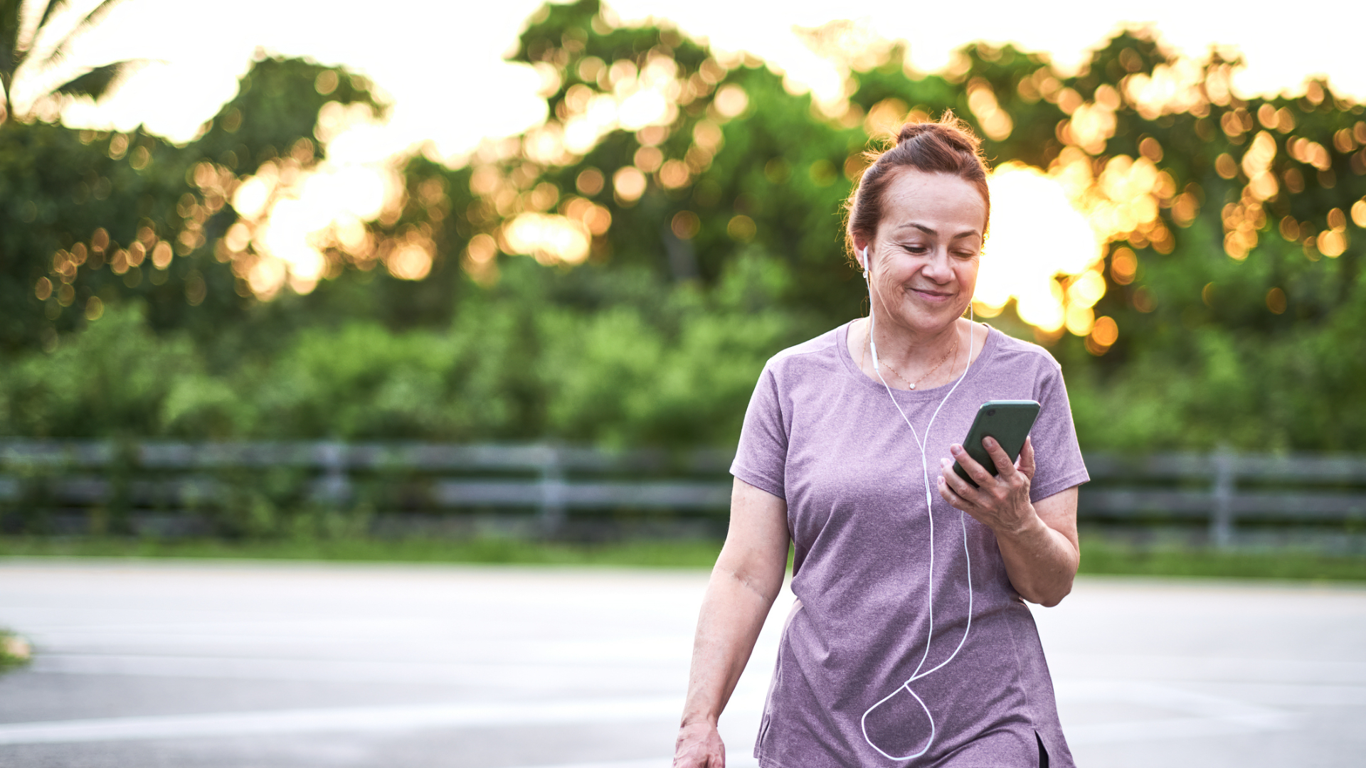 Kvinne som går en tur ute og med høretelefoner i og smiler til smarttelefonen sin.
