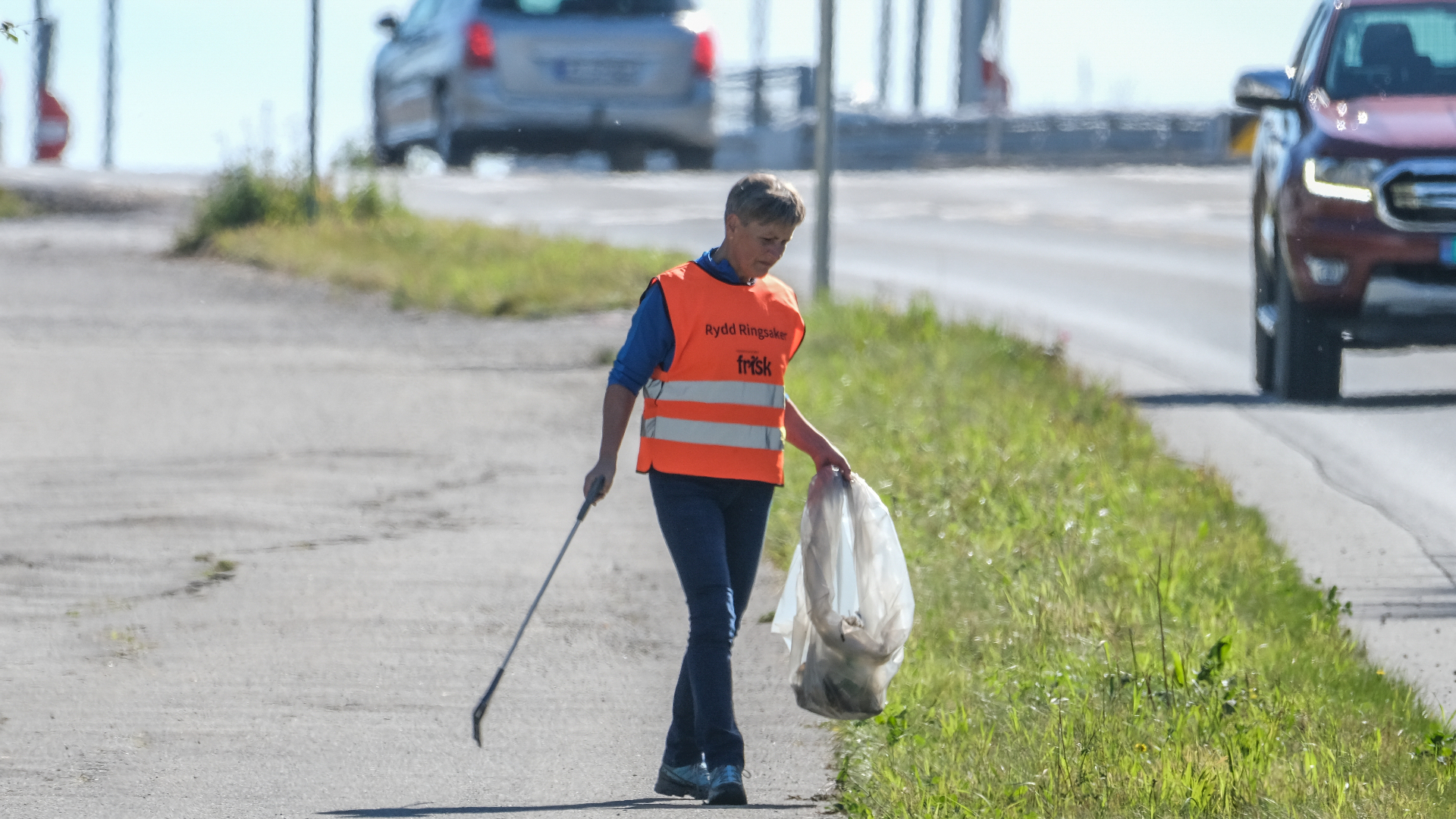 Kvinne med oransj refleksvest som plukker opp søppel langs veien.