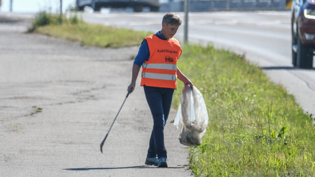 Kvinne med oransj refleksvest som plukker opp søppel langs veien.